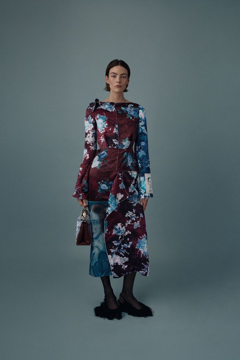 Full-length portrait of an adult female fashion model wearing a floral print dress and holding a purse in a studio setting.
