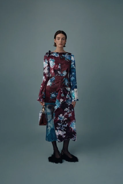 Full-length portrait of an adult female fashion model wearing a floral print dress and holding a purse in a studio setting.