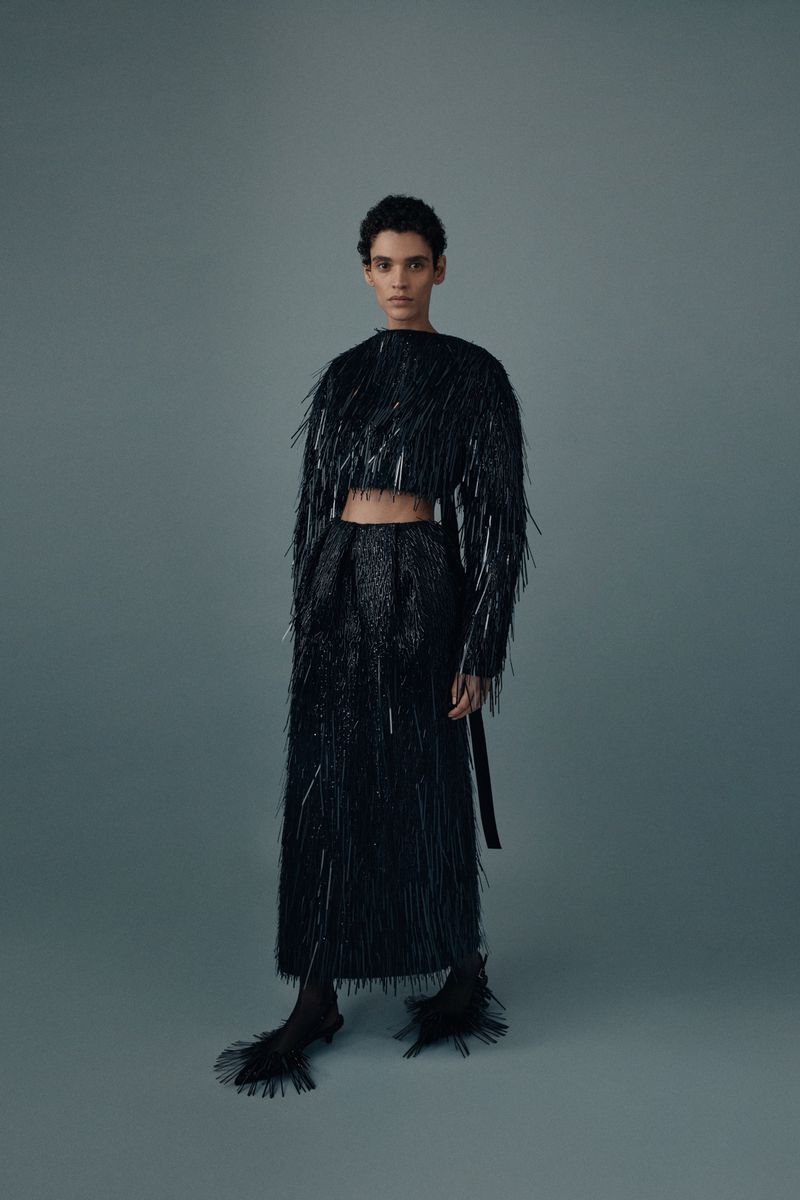 A woman stands in a grey photo studio wearing a black fringe crop top and long skirt with matching black heels, posing for a fashion shot.