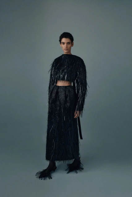 A woman stands in a grey photo studio wearing a black fringe crop top and long skirt with matching black heels, posing for a fashion shot.