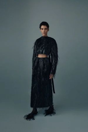 A woman stands in a grey photo studio wearing a black fringe crop top and long skirt with matching black heels, posing for a fashion shot.