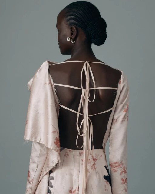 Back view of a slender dark-skinned woman with updo hairstyle wearing an open-back light pink dress with floral pattern against light blue backdrop.
