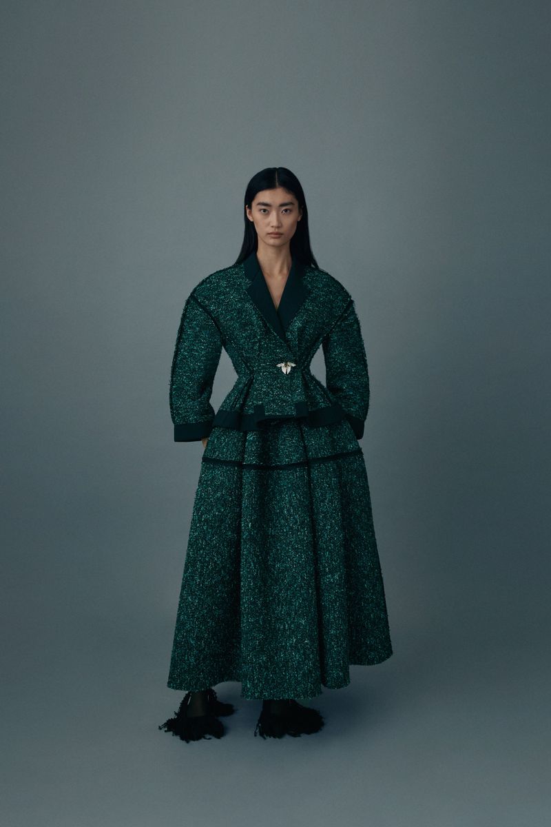 An Asian woman poses in a dark green textured coat and long skirt with black accents against a grey background in a fashion editorial.