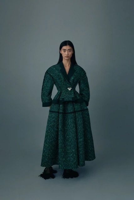 An Asian woman poses in a dark green textured coat and long skirt with black accents against a grey background in a fashion editorial.