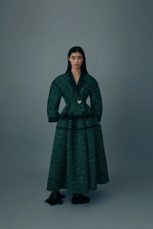 An Asian woman poses in a dark green textured coat and long skirt with black accents against a grey background in a fashion editorial.