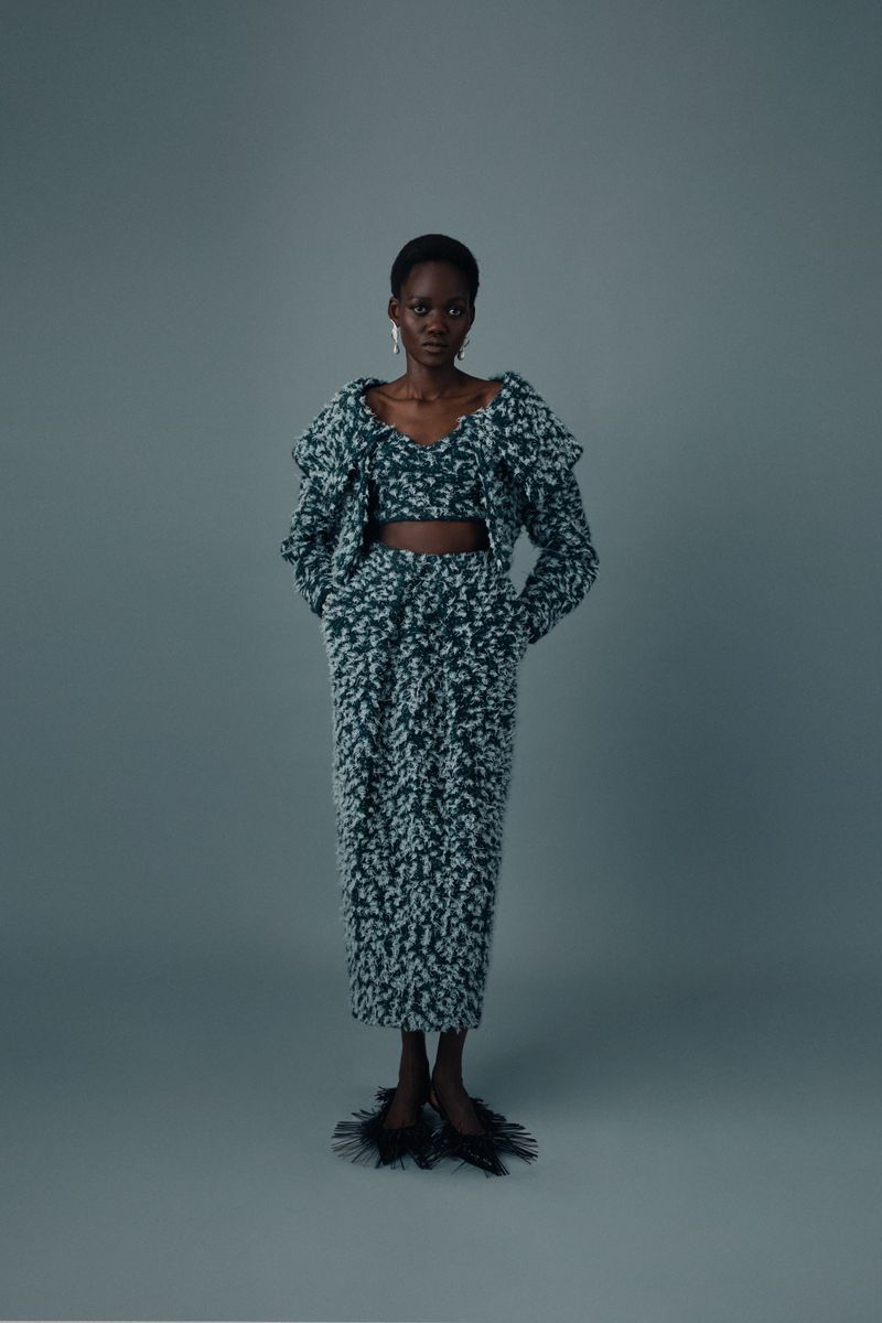 A dark-skinned fashion model poses in a matching green and white knit top, jacket, and skirt on a grey seamless background.