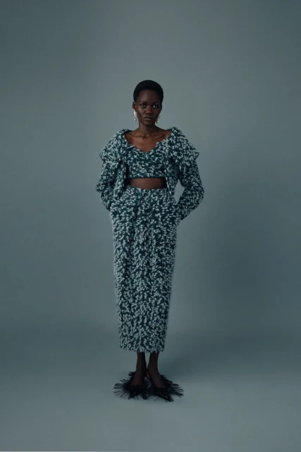 A dark-skinned fashion model poses in a matching green and white knit top, jacket, and skirt on a grey seamless background.