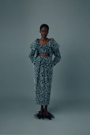 A dark-skinned fashion model poses in a matching green and white knit top, jacket, and skirt on a grey seamless background.