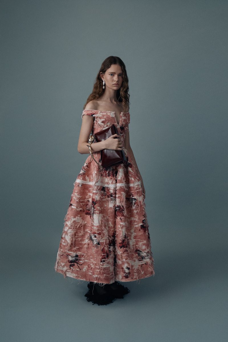 Model wearing a textured off-the-shoulder pink patterned gown and holding a burgundy clutch against a muted grey-blue studio backdrop.