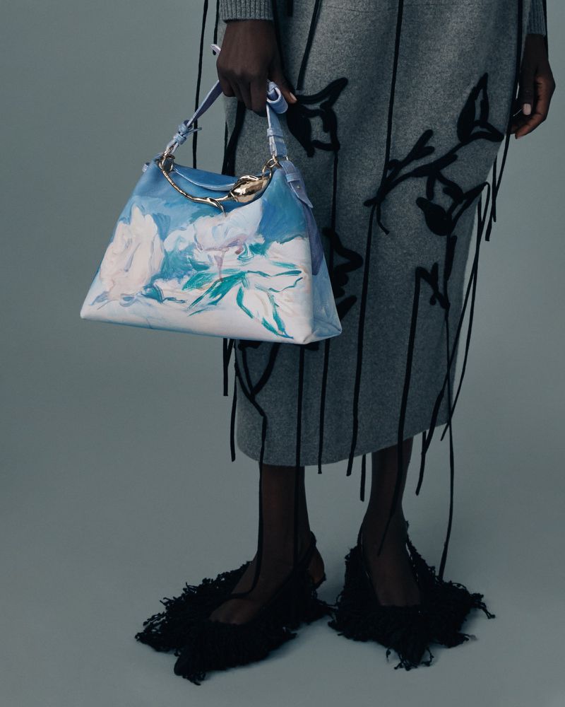 Fashion shot featuring a woman displaying a blue and white floral handbag, paired with a grey skirt and black fringed shoes.