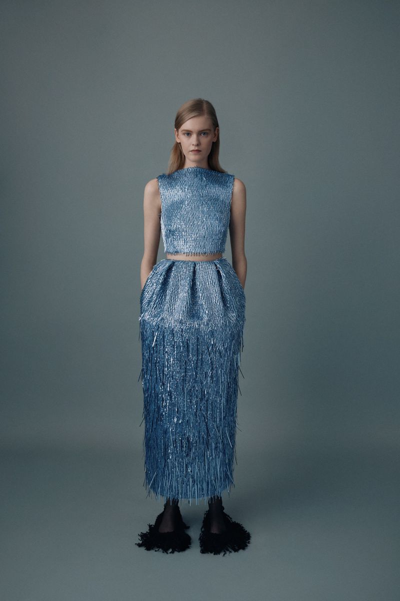 Blond woman in a studio wearing a sparkling blue two-piece fringe dress with black slippers poses against a blue-gray background.