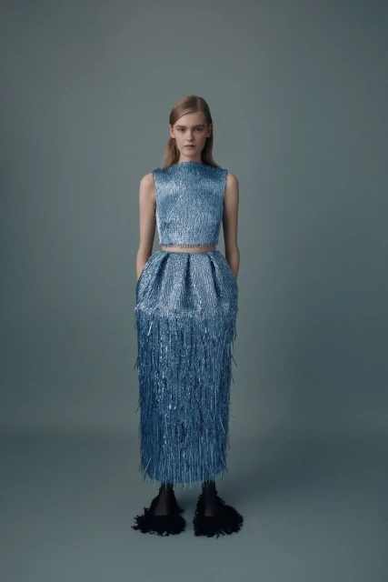 Blond woman in a studio wearing a sparkling blue two-piece fringe dress with black slippers poses against a blue-gray background.