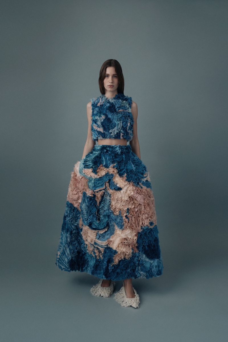 Full-length shot of a fashion model wearing a blue and beige two-piece outfit in a studio with a neutral gray background.