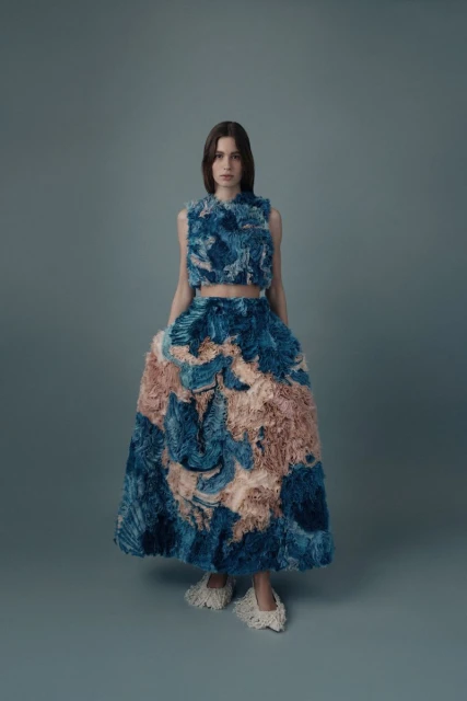 Full-length shot of a fashion model wearing a blue and beige two-piece outfit in a studio with a neutral gray background.