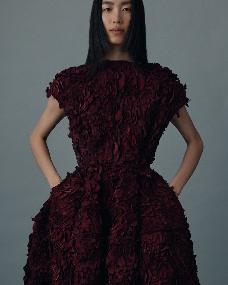 Fashion editorial shot of a single adult female model wearing a burgundy dress with a textured, layered petal design on a grey background.