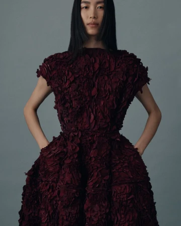 Fashion editorial shot of a single adult female model wearing a burgundy dress with a textured, layered petal design on a grey background.