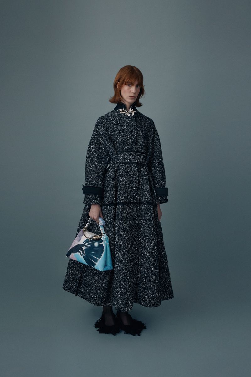 A fashionable adult female in a gray tweed coat and matching skirt holds a patterned handbag in a studio fashion shot.