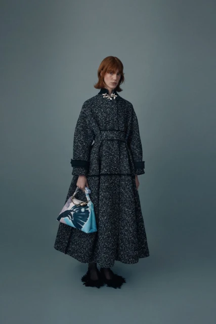 A fashionable adult female in a gray tweed coat and matching skirt holds a patterned handbag in a studio fashion shot.