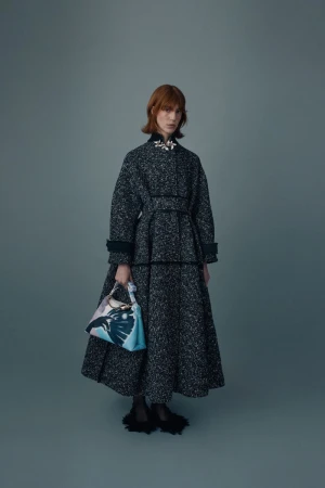 A fashionable adult female in a gray tweed coat and matching skirt holds a patterned handbag in a studio fashion shot.