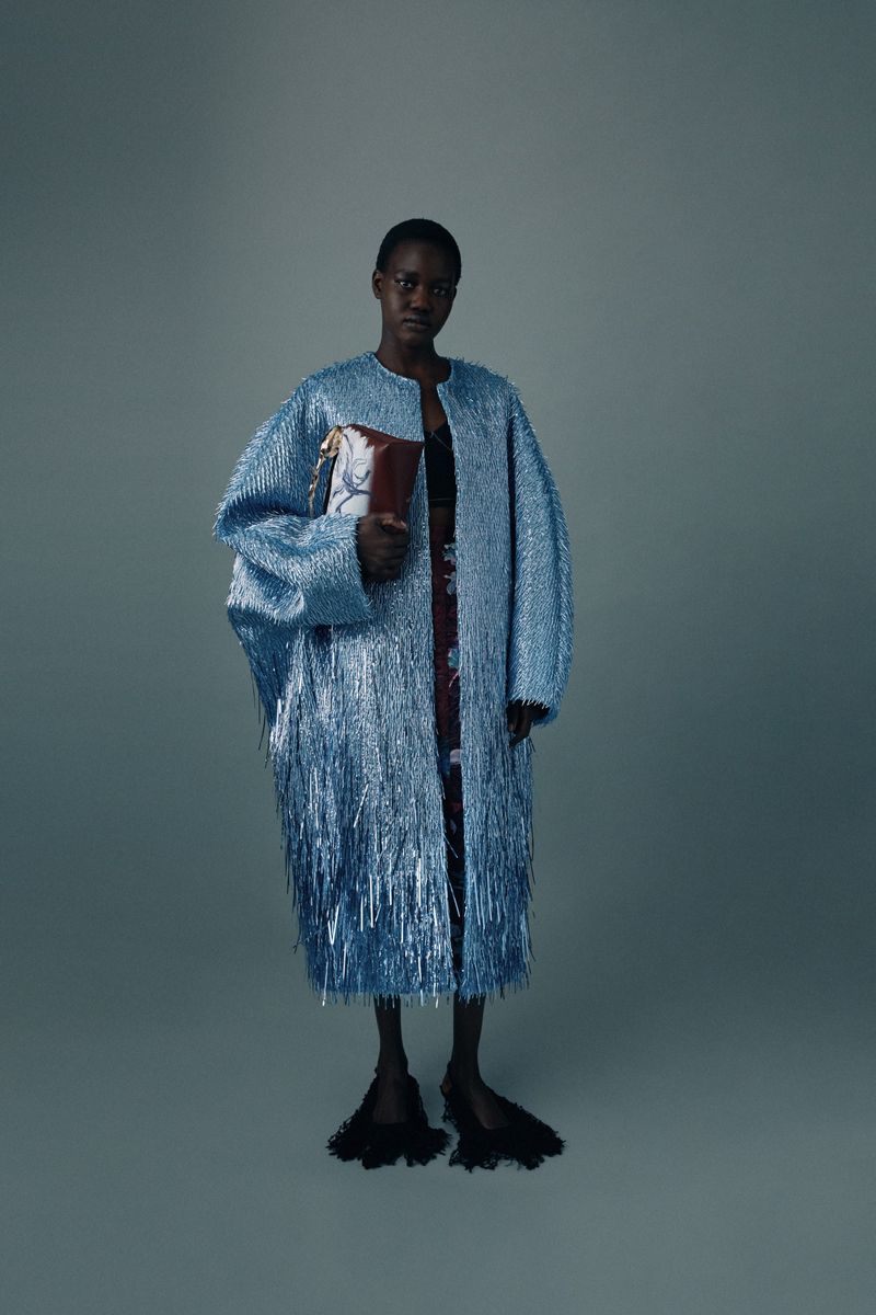 Full-length shot of a Black female model in a light blue, fringed coat, holding a patterned bag, posing in a studio with a grey background.