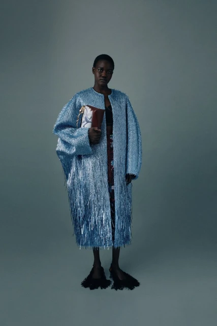 Full-length shot of a Black female model in a light blue, fringed coat, holding a patterned bag, posing in a studio with a grey background.
