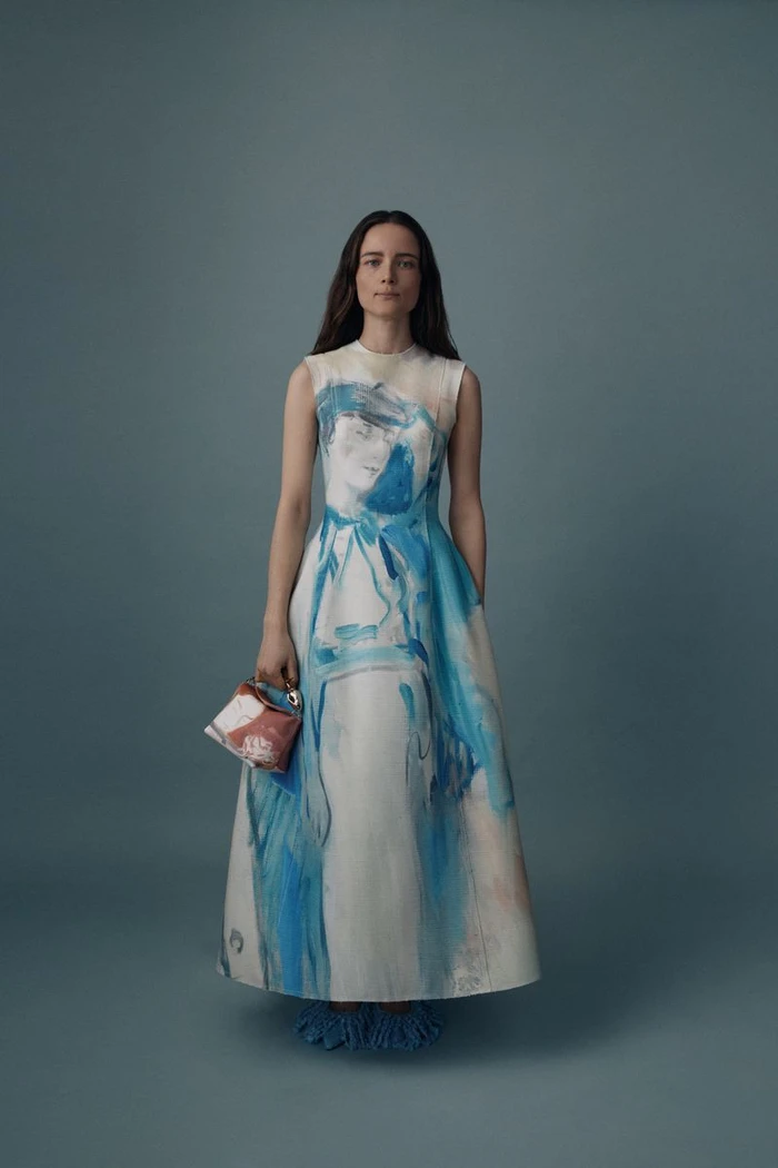 Model wearing a white and blue patterned midi dress holding a small handbag against a solid grey backdrop
