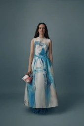 Model wearing a white and blue patterned midi dress holding a small handbag against a solid grey backdrop