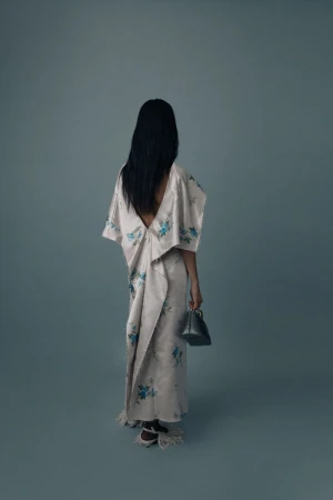 Model seen from behind wearing a draped floral open-back gown and holding a small handbag against a muted blue studio backdrop.