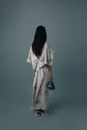 Model seen from behind wearing a draped floral open-back gown and holding a small handbag against a muted blue studio backdrop.