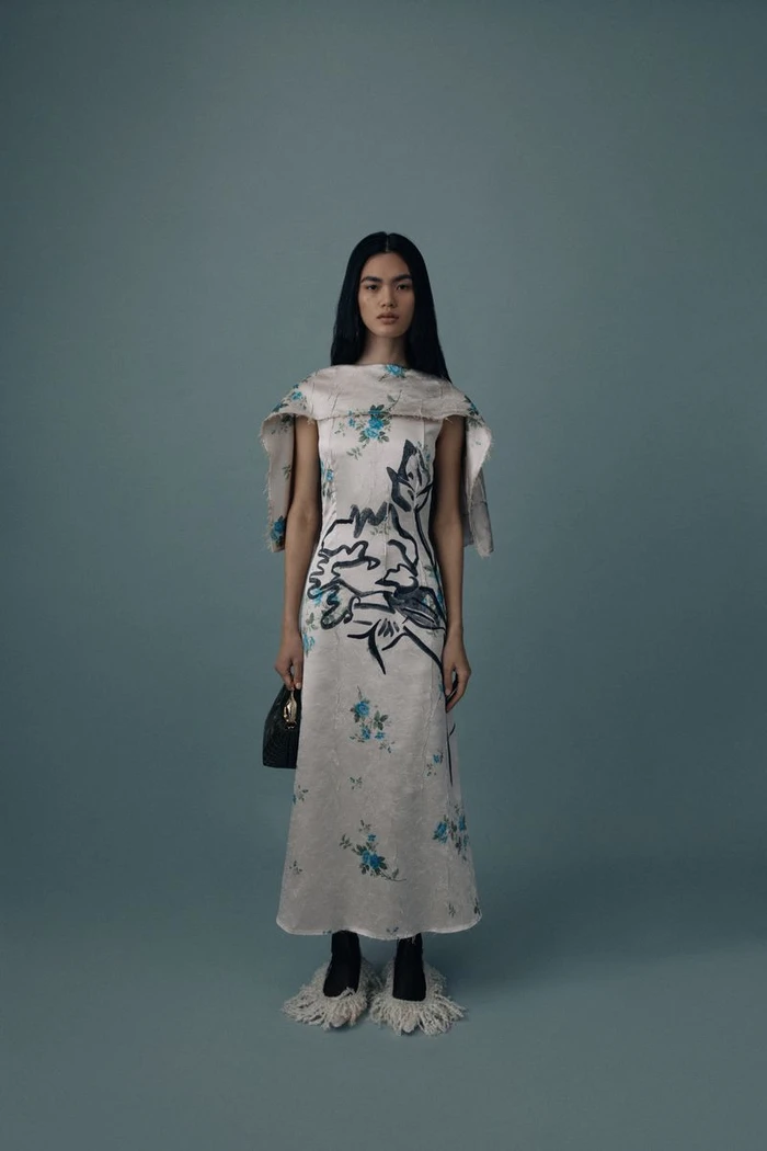 Model wearing a patterned midi dress with a capelet overlay and fringed footwear against a solid grey background