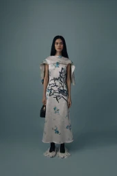 Model wearing a patterned midi dress with a capelet overlay and fringed footwear against a solid grey background