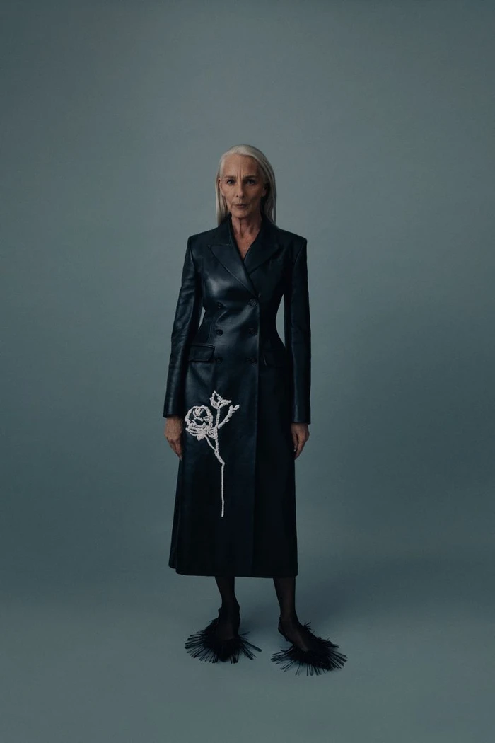 A woman with long grey hair wearing a tailored black leather coat with white floral embroidery against a plain grey-blue studio backdrop.
