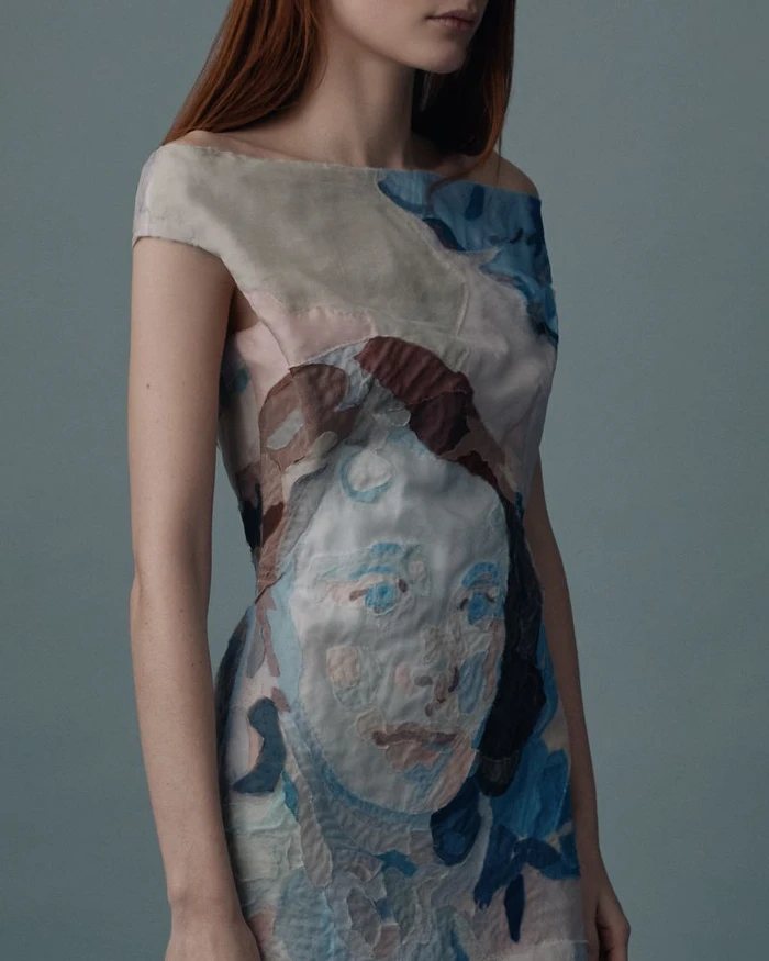 Model wearing an off-the-shoulder dress featuring an artistic textile portrait print against a slate grey studio backdrop.