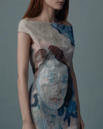 Model wearing an off-the-shoulder dress featuring an artistic textile portrait print against a slate grey studio backdrop.