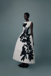 Model wearing a sleeveless ivory bell-shaped gown with black floral applique against a muted grey-blue studio backdrop.