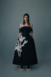 Model wearing a black off-the-shoulder A-line dress with white floral embroidery against a muted blue-grey studio background.