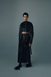 Model posing in a black textured fringed crop top and matching long skirt with feathered heels against a solid grey studio background.