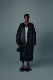 Black woman standing against a grey backdrop wearing a black textured fringed coat holding a small handbag