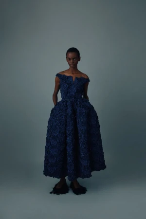 Model wearing an off-the-shoulder textured navy blue flared dress against a muted blue-grey seamless studio backdrop.