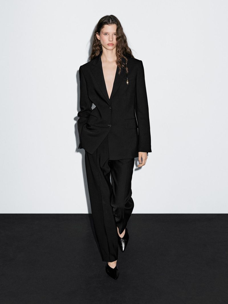 A fashionable adult woman with long wavy hair poses in a black blazer, pants, and shoes against a stark white and black background.