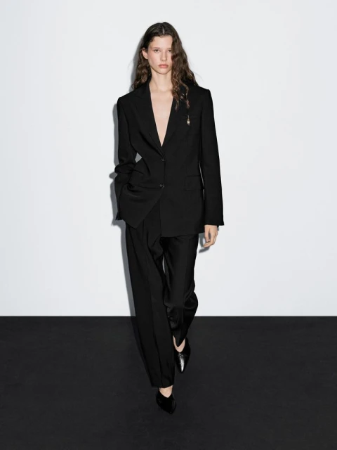 A fashionable adult woman with long wavy hair poses in a black blazer, pants, and shoes against a stark white and black background.