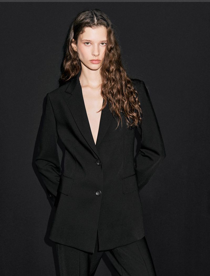 Brunette woman in a black blazer and matching pants poses against a dark backdrop, her gaze directed toward the camera in a fashion shot.