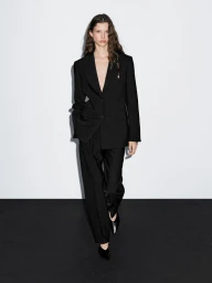 Model in a black suit and trousers standing against a plain white wall with dark flooring.