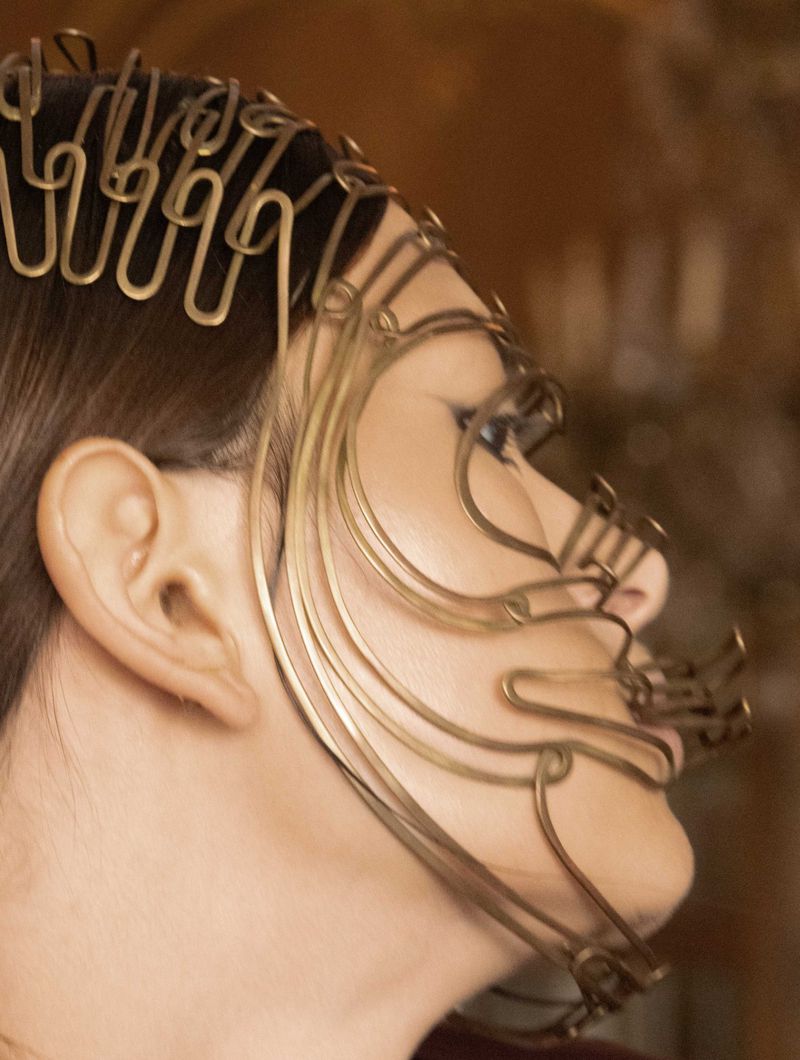 Profile view of an adult female wearing an ornate, gold-colored wire headpiece against a blurred warm-toned background, artistic and stylish.
