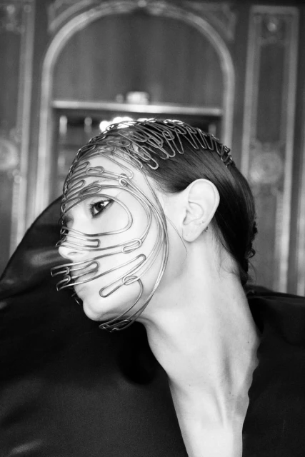 Monochrome portrait of a woman wearing a sculptural mask made of metal wire in an indoor setting, complemented by a high-fashion garment.