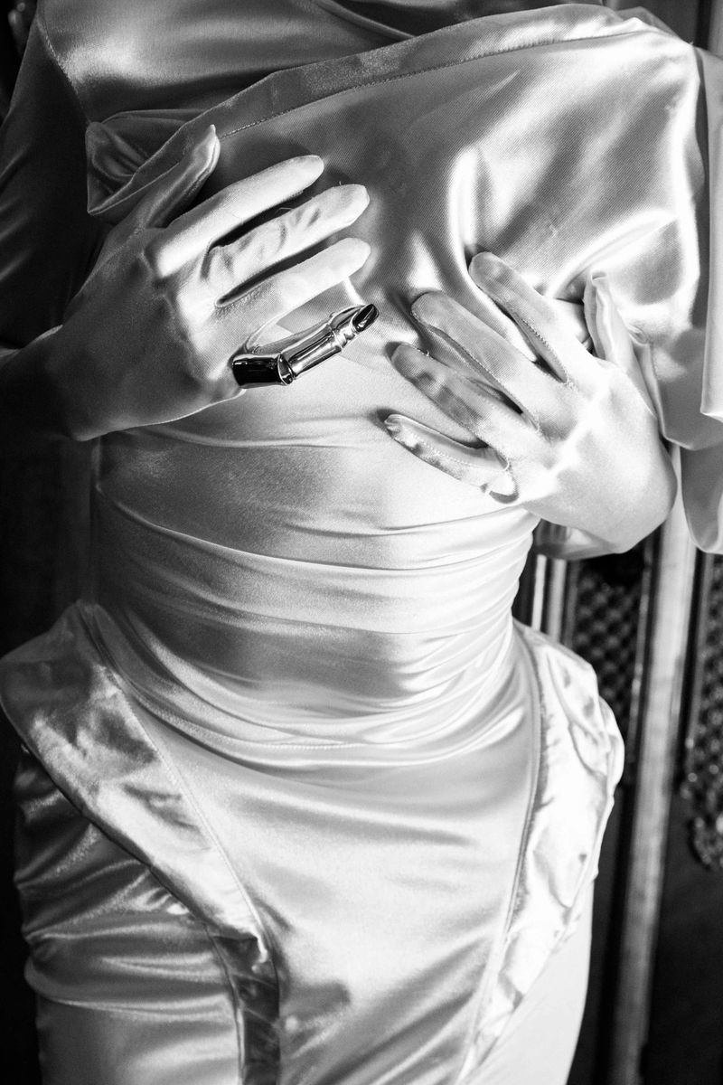 Close-up studio shot in black and white of a person in a silky dress with white gloves holding a lipstick tube against their chest.
