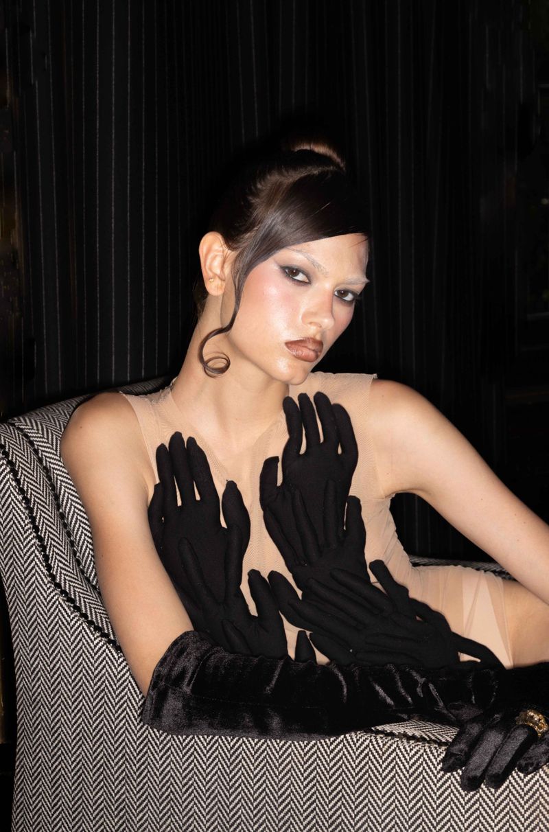 Portrait of a model wearing a nude-colored dress and black gloves while sitting in a herringbone-patterned chair with black pinstripe background.