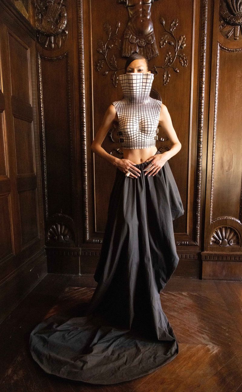 A fashion photo features a woman wearing a unique, high-necked, grid-patterned crop top and a long, flowing dark skirt in an elegant setting.