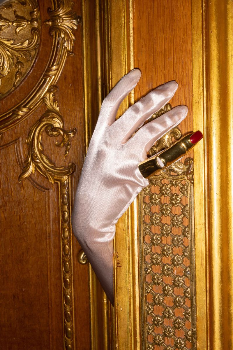 Elegant hand in satin glove holds gold lipstick against a luxurious gold-trimmed wooden panel, evoking classic beauty.