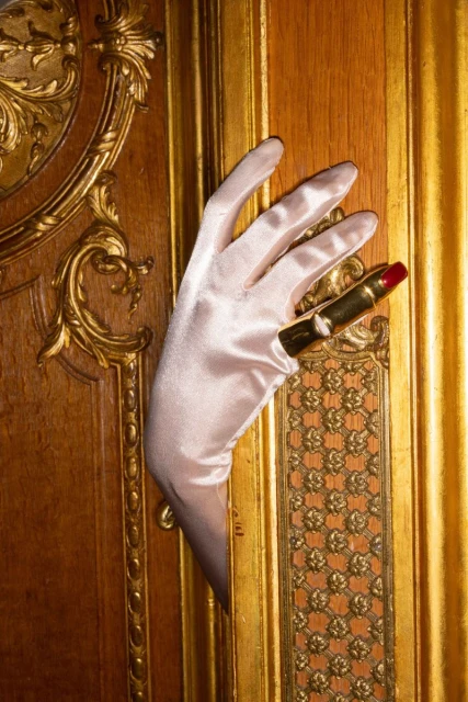 Elegant hand in satin glove holds gold lipstick against a luxurious gold-trimmed wooden panel, evoking classic beauty.